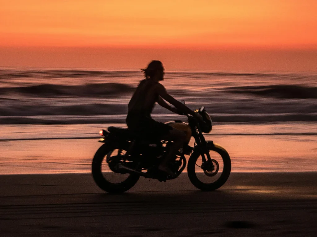 A man riding a motorcycle along the beach shore with the sunset in its background