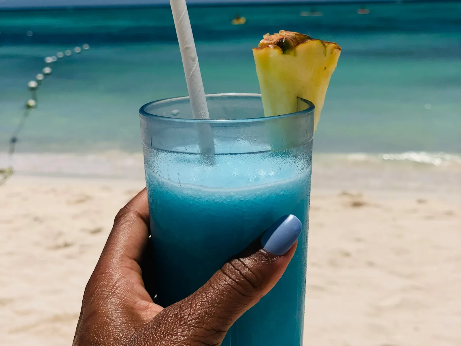 A woman’s hand holding a glass of blue cocktail drink with a white paper straw and a slice of pineapple