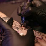 A pair of hands wearing black latex gloves, with the right hand holding a tattoo pen and the left hand holding onto a person's arm while tattooing