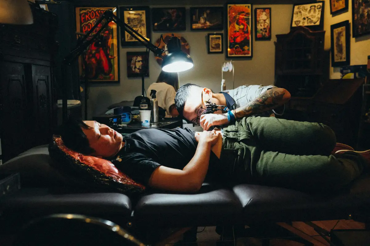A man wearing a black t-shirt is lying down while getting a tattoo on his left hip Image Filename: Best Tattoo Shops & Studios, Krabi