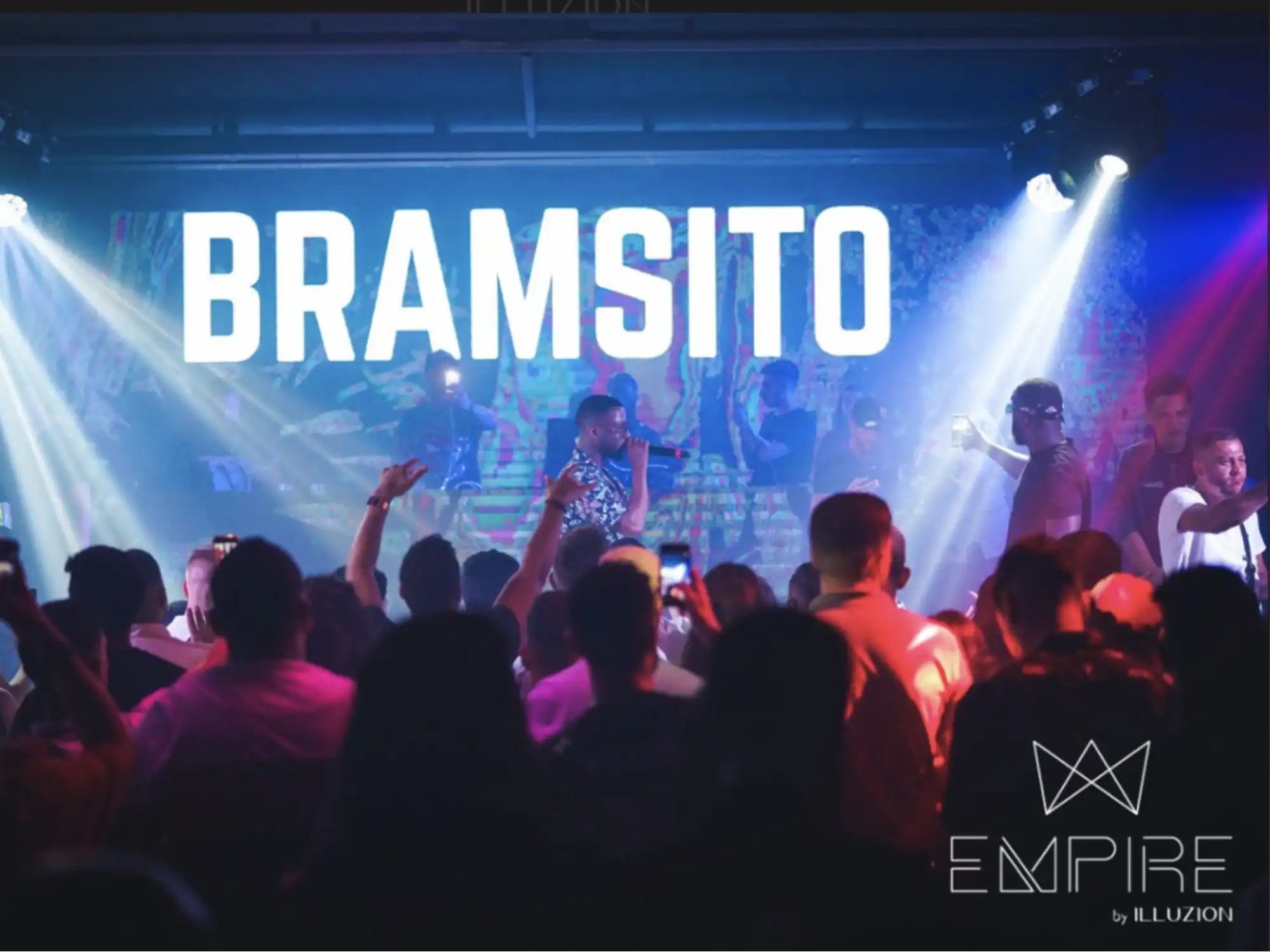 A lively club atmosphere with the word "BRAMSITO" illuminated on stage, with beams of stage lights cutting across the scene. A crowd of people with raised hands appears to be enjoying the performance, encapsulating a high-energy nightlife event at EMPIRE by Illuzion.