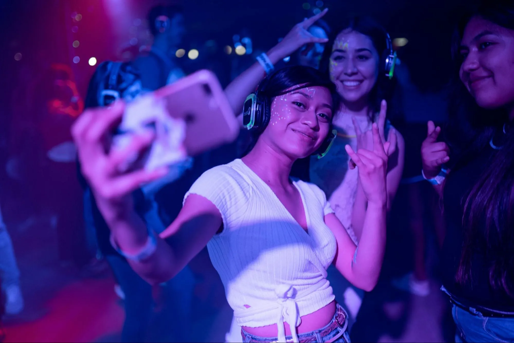 A joyful young woman takes a selfie with friends at White Room Night Club, with vivid blue and purple lighting reflecting their smiling faces and festive glow paint, capturing a moment of fun and connection.
