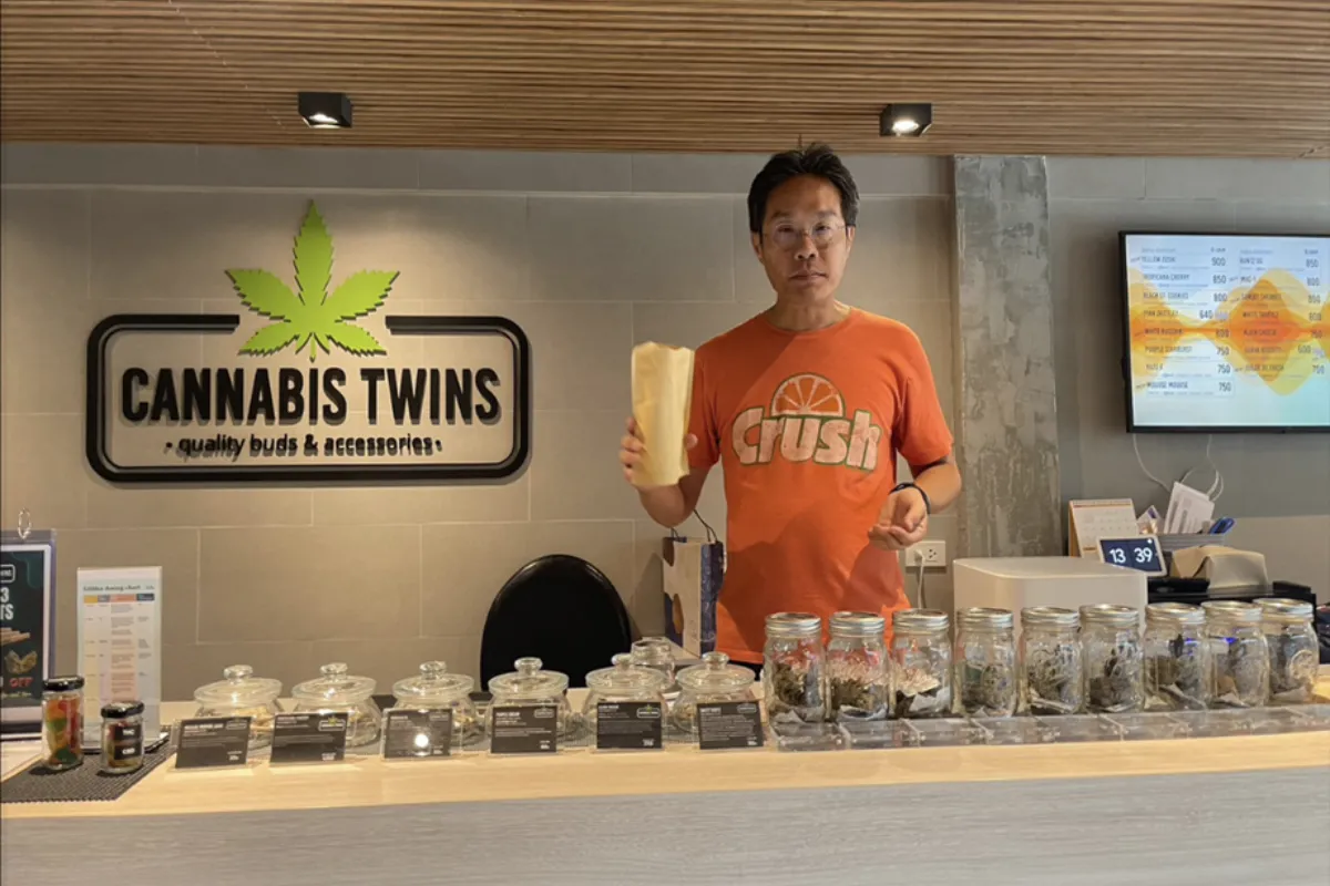 A man wearing eyeglasses and an orange t-shirt is holding a piece of paper bag and is standing in front of the Cannabis Twin’s array of glass jars filled with various weed strains