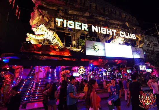 The entrance to Tiger Night Club illuminated at night with vibrant lighting and a large tiger statue overhead. People gather and socialize on the steps and street in front of the club, adding to the lively atmosphere.