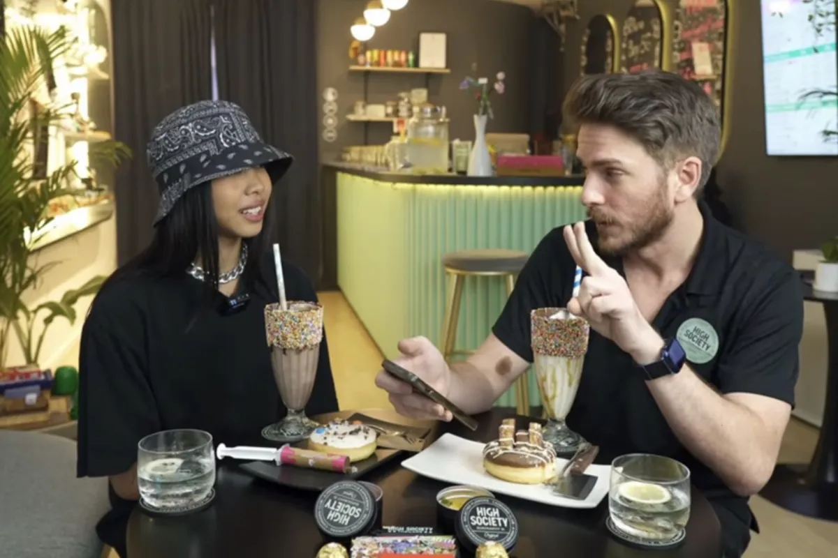 A man and a woman wearing a bucket hat are dining inside High Society Thailand, featuring various cannabis products, doughnuts, and shakes