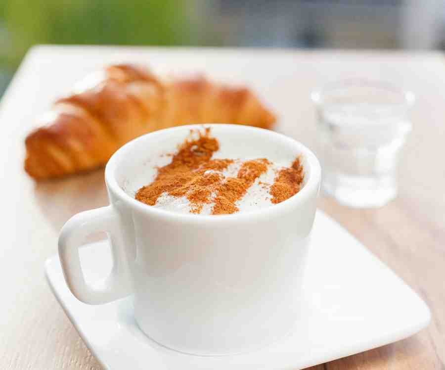 A cup of coffee with croissant and a small glass of water on the background