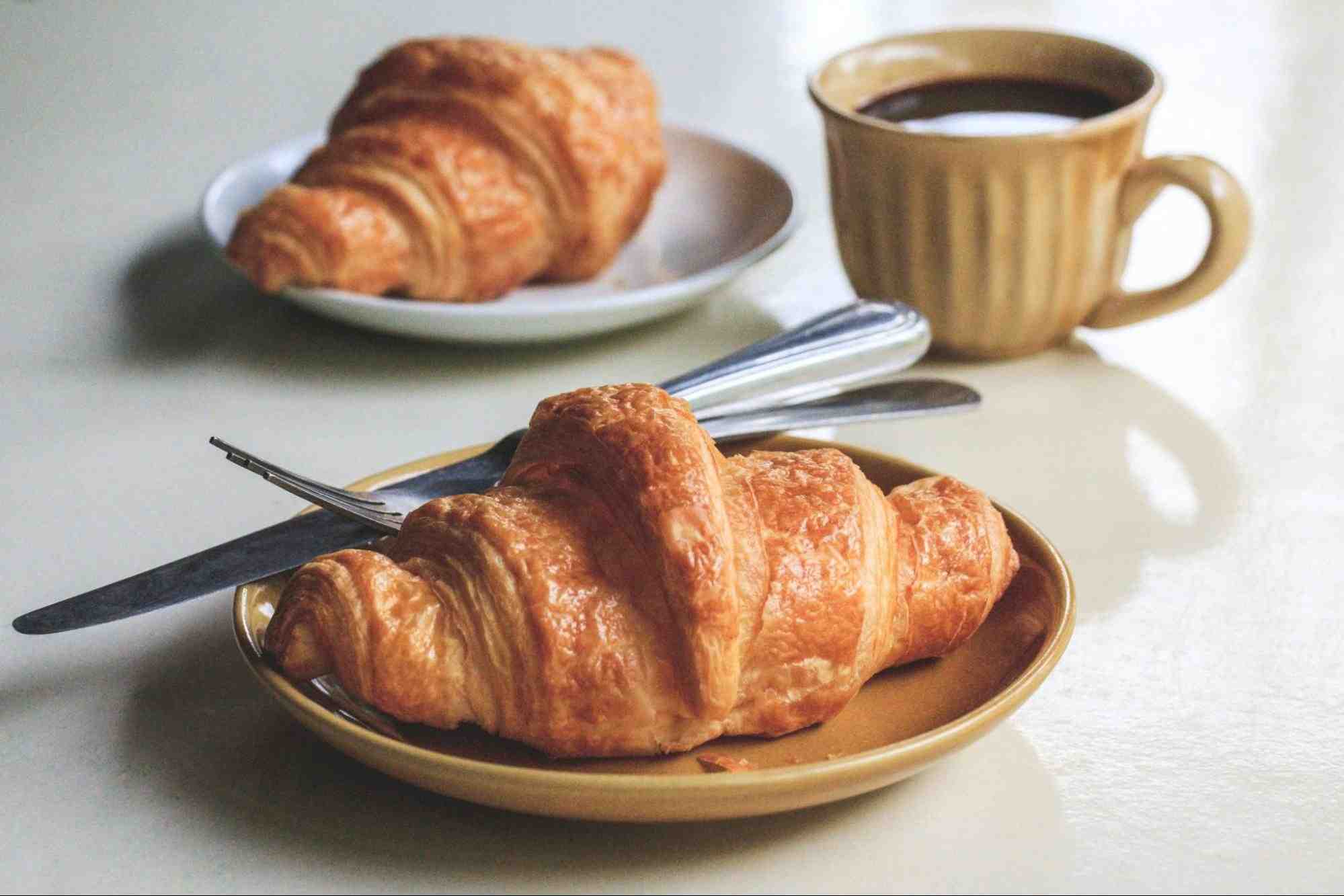 Two small plates of croissants and a cup of black coffee on top of a flat white surface