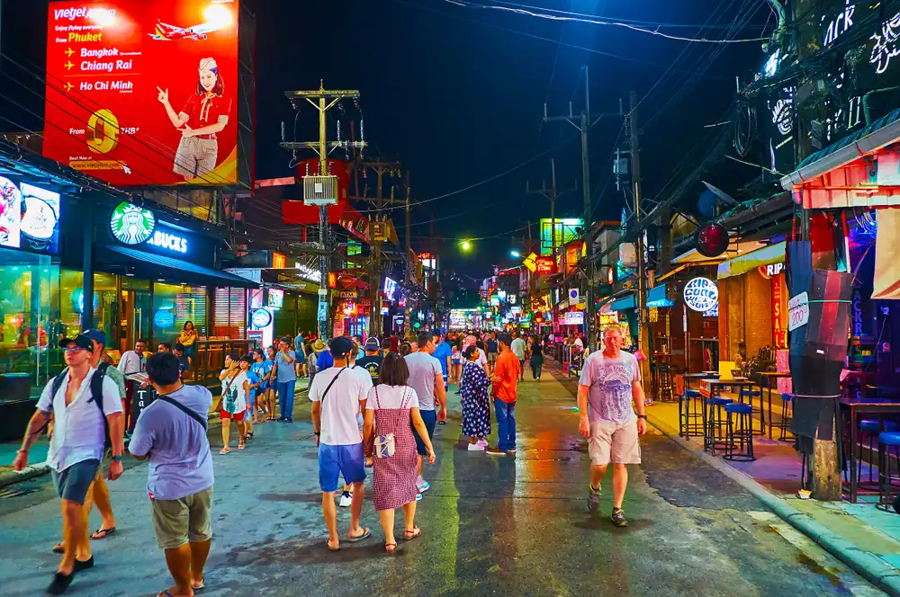 A vibrant and crowded street scene at night on Bangla Road, Phuket, with pedestrians strolling past an array of glowing neon signs and businesses, including a Starbucks. A large advertising board showcasing flight deals adds to the bustling urban atmosphere.