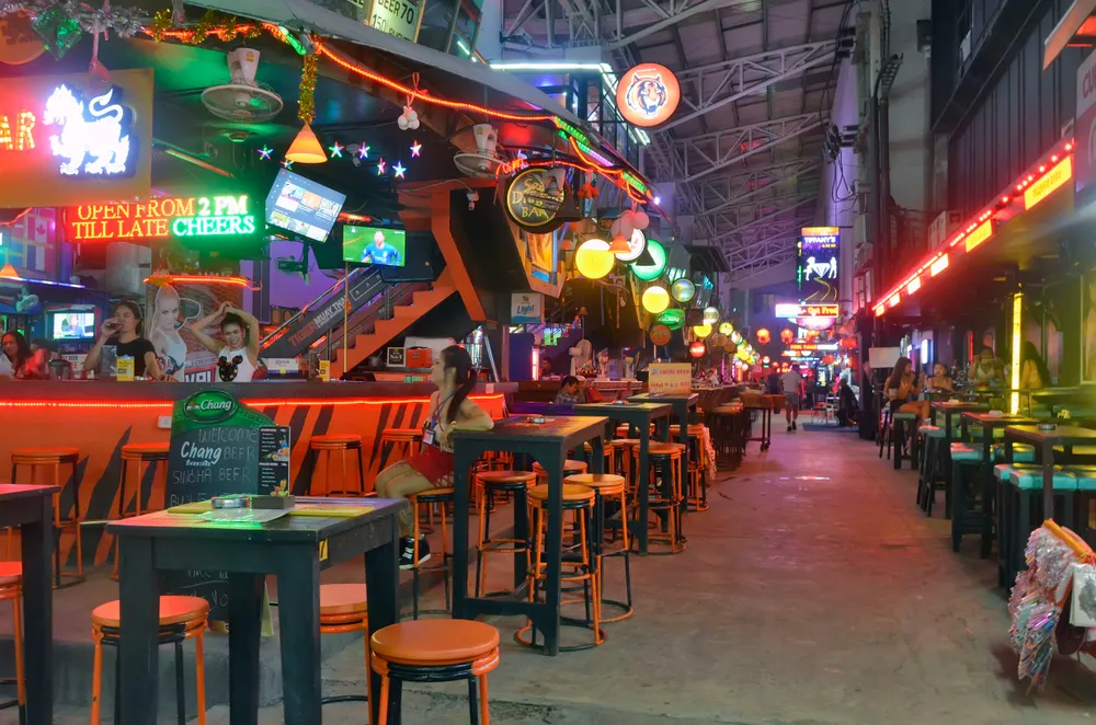 This is an indoor alleyway lined with colorful bar stools and tables, neon signs, and inviting bar fronts on Bangla Road. A few patrons are seen enjoying drinks and conversation in a lively yet relaxed ambiance.