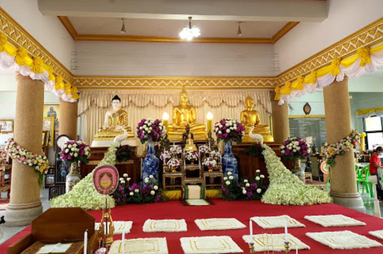 A view of three Buddhist statues with flowers surrounding them and several mats on the floor at Wat Samakkhi Phadungpan in Surat Thani 