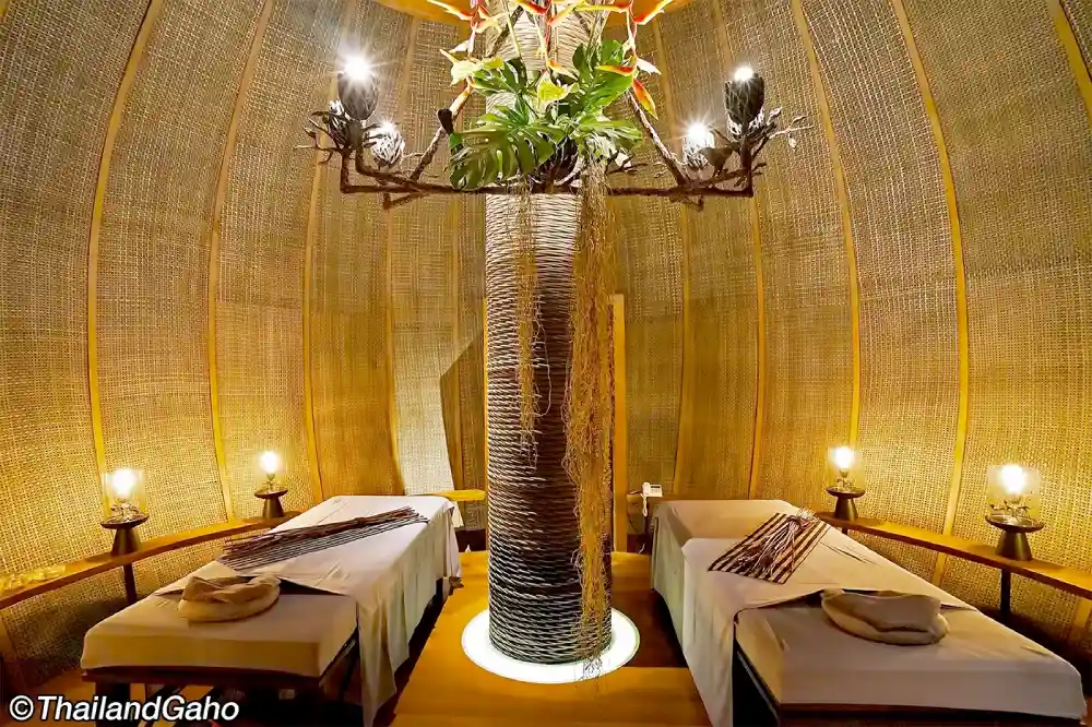 An atmospheric spa room with two massage beds, textured walls, a central tree wrapped in rope with a light installation, and soft lighting creating an ambient and serene environment.