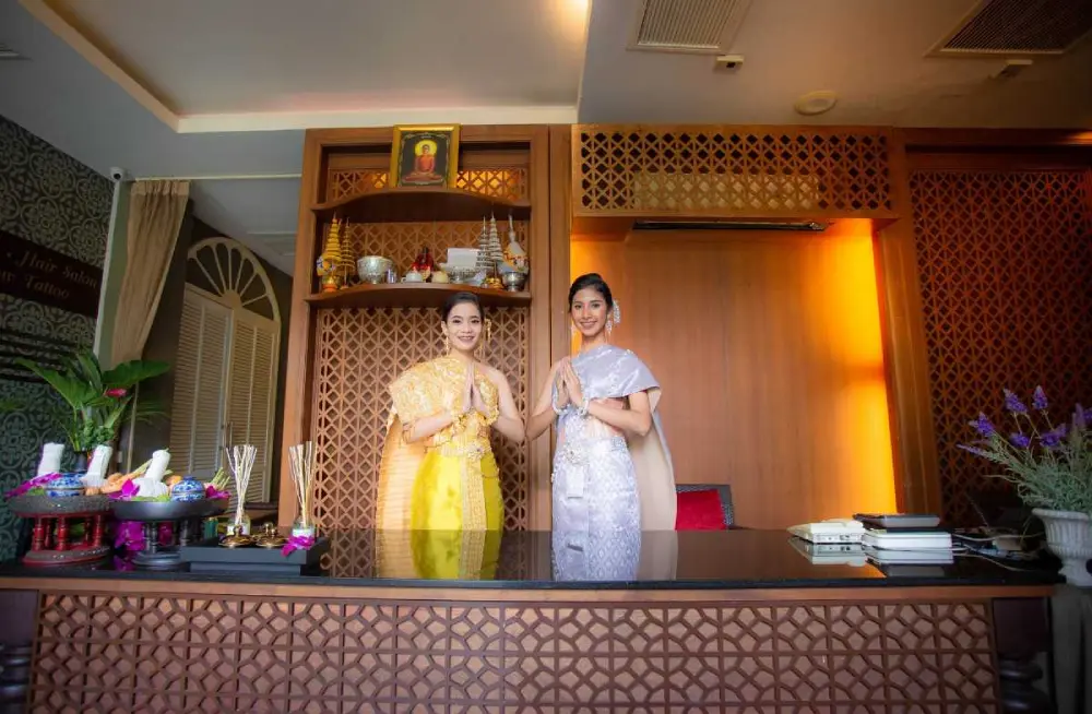 Two welcoming spa staff members, dressed in traditional Thai attire, standing behind the reception desk with a warm and inviting smile in a well-decorated spa lobby.