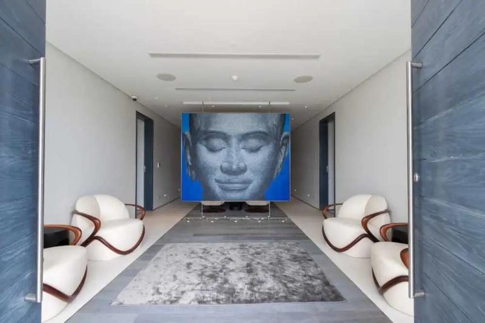 A minimalist and modern lounge area with stylish white chairs, a large grey rug, and a striking blue wall featuring an oversized image of a serene Buddha face.
