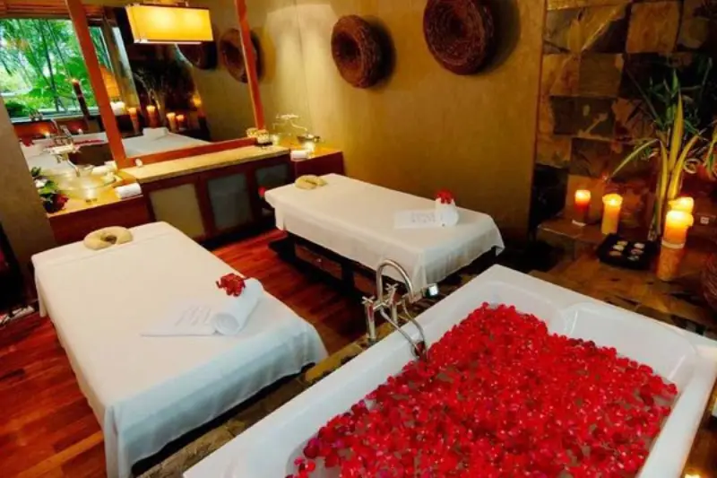 An inviting spa room with two massage tables and a luxurious bath filled with red rose petals, surrounded by candles and warm ambient lighting.