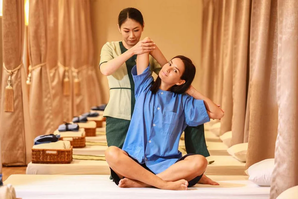 A massage therapist performing a traditional Thai massage on a client seated in a cross-legged position on a mat in a tranquil spa setting with earth-toned curtains and decor.

