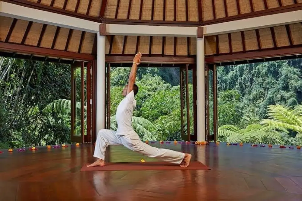 A person practicing yoga in a serene indoor setting with a high ceiling, wooden floors, and large windows overlooking a lush forest.