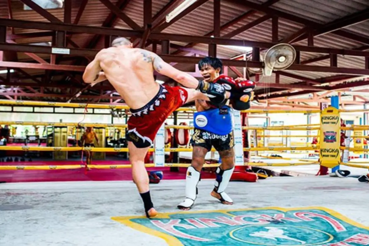 A fighter delivering a high kick to a trainer with pads in Kingka Supa Muay Thai gym.