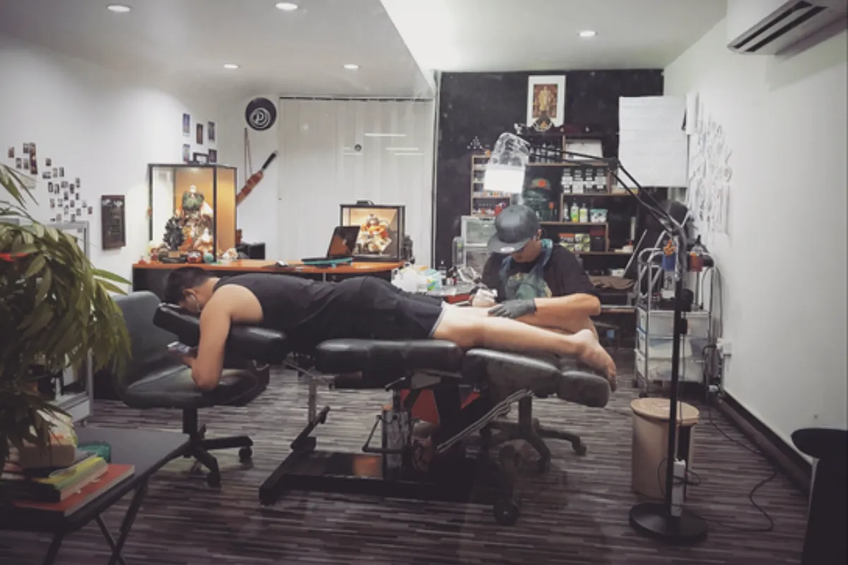 A man lying face down is getting a tattoo in his right leg done by an artist at Jub Tattoo Chiang Mai studio