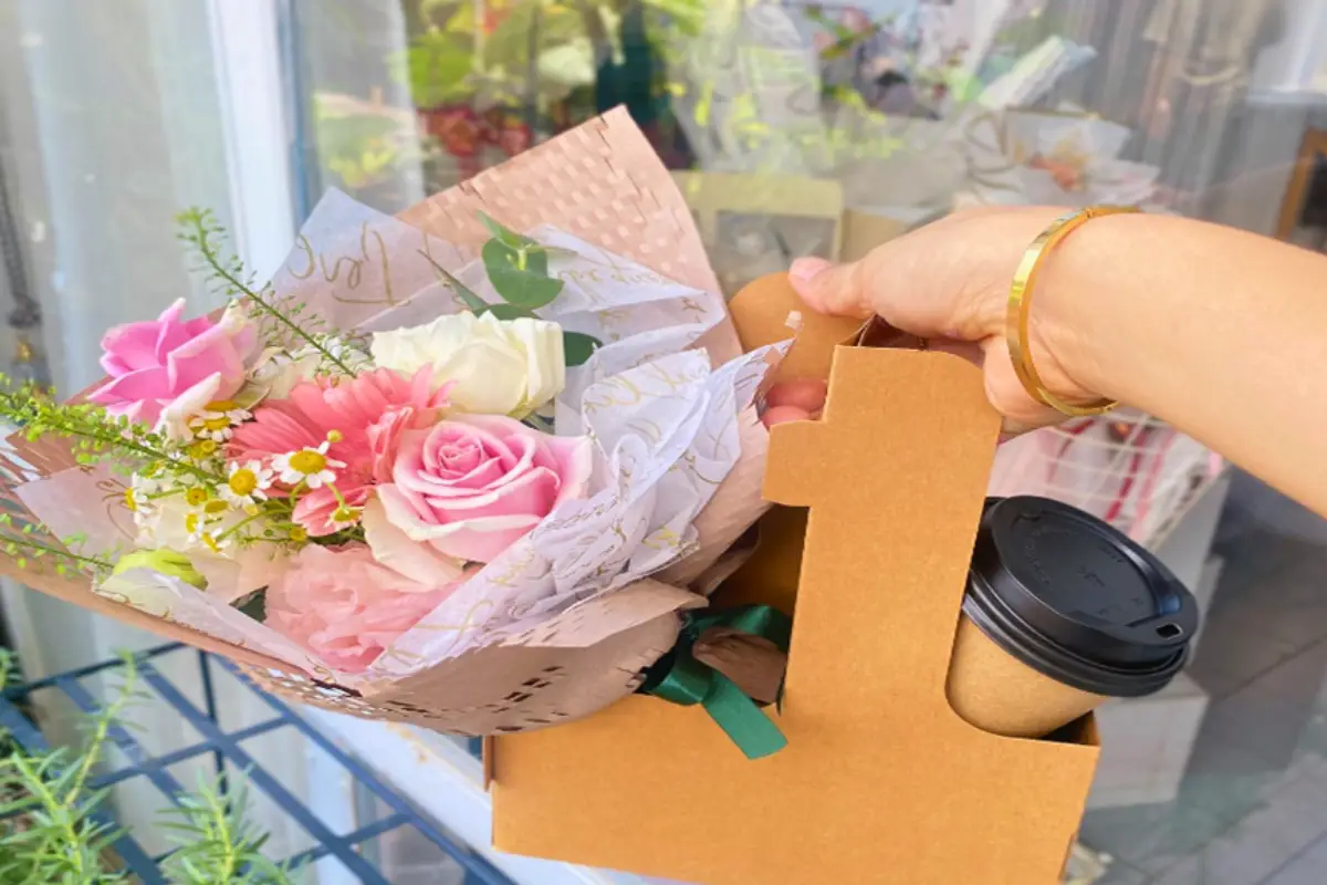 A person is holding a cardboard holder with a fresh flower bouquet and a cup of coffee crafted by Anemone Flower Shop in Chiang Mai